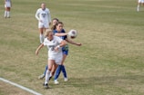 Two female soccer players in action on a field, one wearing a white jersey with 'Hoyas' written on the front and the other in a blue jersey. The player in white is reaching out to control a soccer ball in mid-air. The field is grassy and other players can be seen in the background.