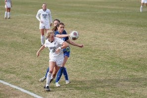 Two female soccer players in action on a field, one wearing a white jersey with 'Hoyas' written on the front and the other in a blue jersey. The player in white is reaching out to control a soccer ball in mid-air. The field is grassy and other players can be seen in the background.