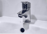 A chrome faucet is mounted on a white sink. A small stream of water is dripping from the tap's spout into the basin below. The background is a plain light color, enhancing the metallic shine of the faucet.