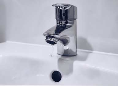 A chrome faucet is mounted on a white sink. A small stream of water is dripping from the tap's spout into the basin below. The background is a plain light color, enhancing the metallic shine of the faucet.