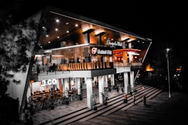 A modern, illuminated building with two levels and a distinctive architectural style. The building contains multiple establishments, including a prominently displayed 'Bakmi GM' restaurant. The structure is surrounded by large glass panels and pillars, with stairs leading up to the entrance. There are people walking around the entrance and the outdoor area is well-lit, highlighting the contemporary design elements.