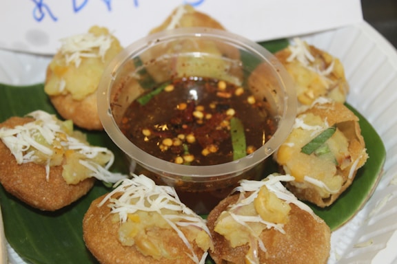 A plate featuring several round, crispy snacks filled with a potato mixture and garnished with white shredded cheese. In the center, there is a small plastic container filled with a reddish-brown sauce containing visible spices and chili flakes. The snacks are arranged neatly on a large green leaf.
