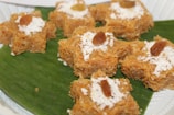 Seven pieces of a brown-colored dessert made from vermicelli or a similar ingredient are arranged on a large green leaf. Each dessert piece is topped with a sprinkle of grated white coconut and has a single raisin in the center.