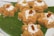 Seven pieces of a brown-colored dessert made from vermicelli or a similar ingredient are arranged on a large green leaf. Each dessert piece is topped with a sprinkle of grated white coconut and has a single raisin in the center.