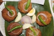 Five golden-brown, crispy cutlets are arranged on a green banana leaf. Each cutlet is topped with a slice of raw onion and a whole green chili. Slices of hard-boiled eggs and additional raw onion are placed in the center for garnish.