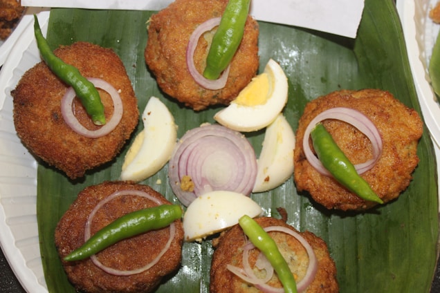 Five golden-brown, crispy cutlets are arranged on a green banana leaf. Each cutlet is topped with a slice of raw onion and a whole green chili. Slices of hard-boiled eggs and additional raw onion are placed in the center for garnish.