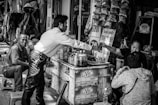 A bustling street-side stall in an urban setting features a man in a white shirt reaching over to hand money to a vendor. The vendor is surrounded by various products including bottles and packets hanging above. Two other individuals are nearby, one sitting on a stool and another with their back turned. The stall is busy with a lot of items displayed, creating a lively marketplace atmosphere.