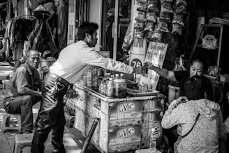 A bustling street-side stall in an urban setting features a man in a white shirt reaching over to hand money to a vendor. The vendor is surrounded by various products including bottles and packets hanging above. Two other individuals are nearby, one sitting on a stool and another with their back turned. The stall is busy with a lot of items displayed, creating a lively marketplace atmosphere.
