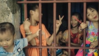 A group of children are interacting playfully behind red metal bars. They are smiling, making peace signs, and holding objects like bracelets and a colorful bag. The background is a dimly lit space, adding contrast to their bright clothing.