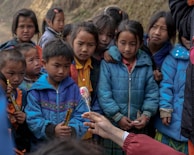 A group of children is gathered around, mostly wearing colorful winter jackets. They appear attentive and curious, looking at a hand holding a lollipop and another cylindrical object.