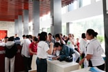 A group of people is gathered around a registration table indoors. Some are leaning over the table filling out forms, while others are standing and conversing. The setting is a well-lit area with natural light coming through large windows. The ceiling has distinctive wooden slats and there are multiple stone pillars.