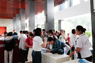 A group of people is gathered around a registration table indoors. Some are leaning over the table filling out forms, while others are standing and conversing. The setting is a well-lit area with natural light coming through large windows. The ceiling has distinctive wooden slats and there are multiple stone pillars.