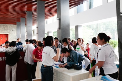 A group of people is gathered around a registration table indoors. Some are leaning over the table filling out forms, while others are standing and conversing. The setting is a well-lit area with natural light coming through large windows. The ceiling has distinctive wooden slats and there are multiple stone pillars.
