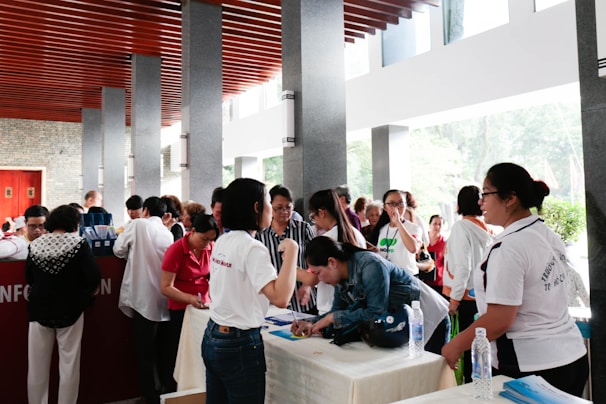 A group of people is gathered around a registration table indoors. Some are leaning over the table filling out forms, while others are standing and conversing. The setting is a well-lit area with natural light coming through large windows. The ceiling has distinctive wooden slats and there are multiple stone pillars.