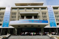 A modern building with geometric patterns on the facade, featuring large vertical banners on each side promoting an event related to World Hepatitis Day. The entrance is bustling with people gathered on the steps leading inside. The banners include several logos and text in a foreign language.