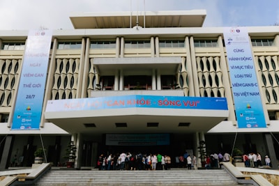 A modern building with geometric patterns on the facade, featuring large vertical banners on each side promoting an event related to World Hepatitis Day. The entrance is bustling with people gathered on the steps leading inside. The banners include several logos and text in a foreign language.