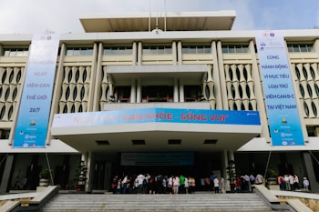 A modern building with geometric patterns on the facade, featuring large vertical banners on each side promoting an event related to World Hepatitis Day. The entrance is bustling with people gathered on the steps leading inside. The banners include several logos and text in a foreign language.