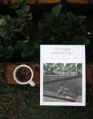 A white magazine titled 'design anthology' is placed on a rustic orange-brown brick surface with green leaves and plants around it. The magazine cover features a photograph of greenery and architecture. Next to the magazine, there is a white cup filled with black coffee.