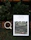 A white magazine titled 'design anthology' is placed on a rustic orange-brown brick surface with green leaves and plants around it. The magazine cover features a photograph of greenery and architecture. Next to the magazine, there is a white cup filled with black coffee.