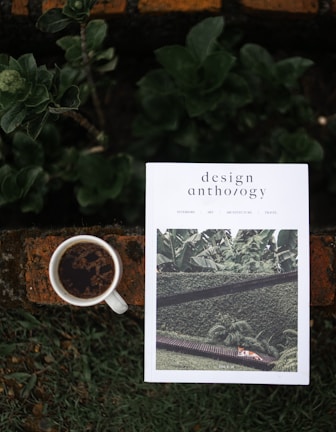 A white magazine titled 'design anthology' is placed on a rustic orange-brown brick surface with green leaves and plants around it. The magazine cover features a photograph of greenery and architecture. Next to the magazine, there is a white cup filled with black coffee.