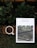 A white magazine titled 'design anthology' is placed on a rustic orange-brown brick surface with green leaves and plants around it. The magazine cover features a photograph of greenery and architecture. Next to the magazine, there is a white cup filled with black coffee.