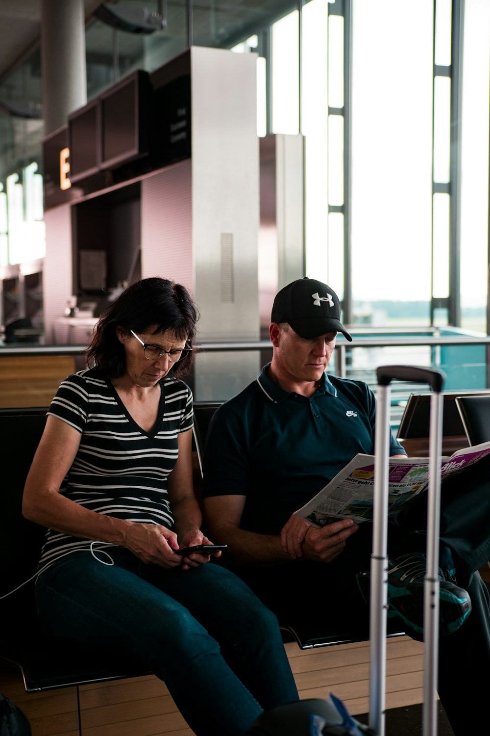 woman fiddling on smartphone beside man with cap reading newspaper