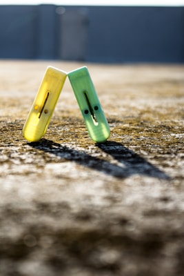 Two small plastic balance toys with smiley faces, one yellow and one green, are positioned on a textured concrete surface, casting long shadows in bright sunlight. They are aligned to form an inverted V shape with a blurred background.