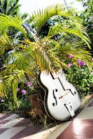 A large, black acoustic guitar is placed outdoors amongst lush green palm fronds and vibrant pink flowers. The setting is bright, with sunlight casting shadows and highlights on the guitar and surrounding foliage. The guitar leans against a stone or brick structure on a tiled floor, adding an artistic contrast to the natural and constructed elements.