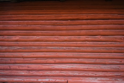 A close-up view of a wooden log wall with a reddish-brown stain, showing the texture and grain of the wood.