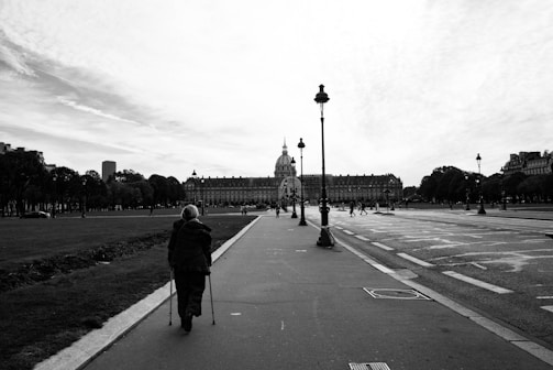 A person walking down a wide, paved path in an urban park setting, using a pair of crutches. The path is lined with grass on one side and a road with clear lane markings on the other. In the background, a large historic building with a dome is visible, surrounded by neatly arranged rows of trees. There are street lamps along the path and a few people in the distance.
