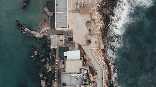 An aerial view of a coastal structure surrounded by the sea. The image captures a rugged shoreline with waves crashing against rocks. The complex consists of several buildings with flat roofs, interconnected by pathways. The ocean is a deep blue-green, contrasting with the light sandy and rocky textures of the land.