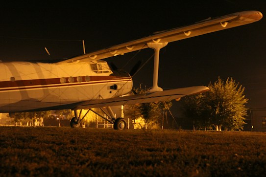 An old-fashioned biplane is parked on a grassy area under a dark night sky. The plane is illuminated by warm yellow lighting, highlighting its side and giving it a dramatic, vintage appearance. Trees and other shrubbery are faintly visible in the background, adding a sense of depth and mystery.