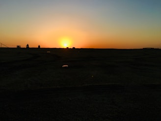 A horizon with a vibrant sunset, casting an orange and yellow glow across the sky. The foreground is a dark silhouette of land with a flat texture, while a couple of small, indistinct figures are visible near the horizon.