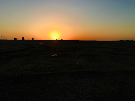 A horizon with a vibrant sunset, casting an orange and yellow glow across the sky. The foreground is a dark silhouette of land with a flat texture, while a couple of small, indistinct figures are visible near the horizon.
