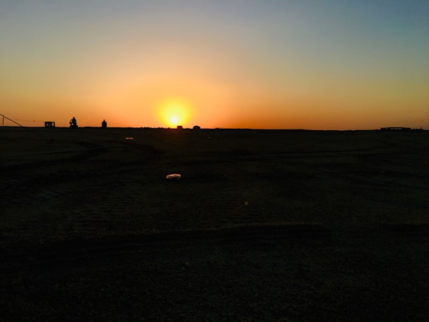 A horizon with a vibrant sunset, casting an orange and yellow glow across the sky. The foreground is a dark silhouette of land with a flat texture, while a couple of small, indistinct figures are visible near the horizon.
