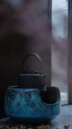 A rustic black teapot sits inside a large, blue ceramic bowl with intricate wave-like patterns. The background reveals a calm, softly lit room with a window partially visible, casting gentle reflections and shadows. Fallen leaves can be seen scattered at the base of the objects, adding an organic touch.