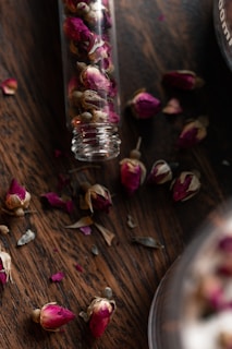 Dried rosebuds are scattered on a wooden surface, with some placed inside a small, transparent glass vial. The pink and purple hues of the rosebuds contrast starkly against the dark wood, creating a rustic and natural arrangement.