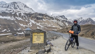 A cyclist stands next to a milestone at Baralacha La Pass, surrounded by snow-capped mountains and a winding road. The sky is overcast, adding a dramatic effect to the rugged landscape.