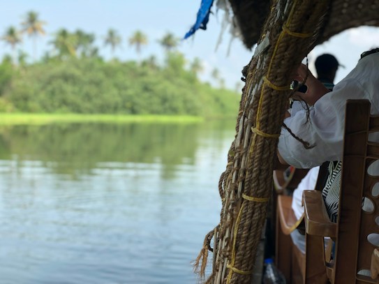 A tranquil river scene with lush green foliage in the background and a boat in the foreground. The boat appears to be made of wood with a canopy woven from ropes. Passengers can be seen on the right side, one holding onto the structure, wearing a white garment.