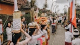 A traditional cultural procession with participants dressed in colorful and ceremonial attire. The scene includes women carrying offerings on their heads and several people gathered around, suggesting a festive or religious event. Decorative elements like flags and bamboo poles line the pathway.