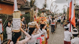 A traditional cultural procession with participants dressed in colorful and ceremonial attire. The scene includes women carrying offerings on their heads and several people gathered around, suggesting a festive or religious event. Decorative elements like flags and bamboo poles line the pathway.