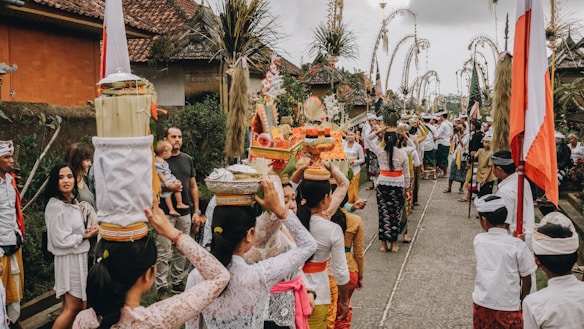 A traditional cultural procession with participants dressed in colorful and ceremonial attire. The scene includes women carrying offerings on their heads and several people gathered around, suggesting a festive or religious event. Decorative elements like flags and bamboo poles line the pathway.