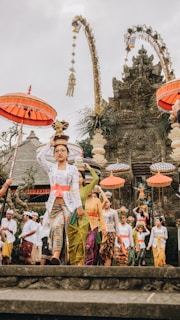 A group of people dressed in traditional Balinese attire participate in a ceremonial procession in front of a richly decorated temple. The procession includes individuals carrying offerings on their heads and is accompanied by ornate umbrellas and colorful garments. The temple facade is intricately carved, contributing to the cultural atmosphere.