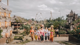 A group of people dressed in traditional Balinese attire walk through a temple complex. The women are carrying baskets on their heads and are surrounded by lush greenery and traditional Balinese architecture. The sky is overcast, adding a serene atmosphere to the scene.