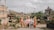 A group of people dressed in traditional Balinese attire walk through a temple complex. The women are carrying baskets on their heads and are surrounded by lush greenery and traditional Balinese architecture. The sky is overcast, adding a serene atmosphere to the scene.