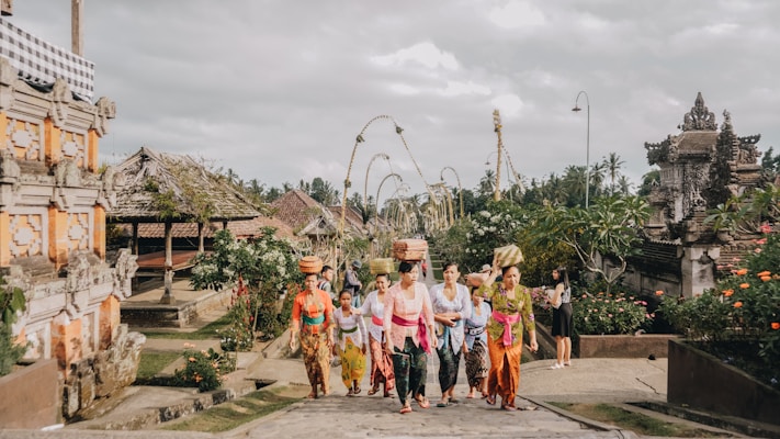 A group of people dressed in traditional Balinese attire walk through a temple complex. The women are carrying baskets on their heads and are surrounded by lush greenery and traditional Balinese architecture. The sky is overcast, adding a serene atmosphere to the scene.