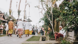 People dressed in traditional clothing walk down a street lined with decorated tall bamboo poles, while an elderly person sits on the side surrounded by greenery.