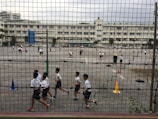 Children are playing and running on a large outdoor playground enclosed by a mesh fence. Various activities are taking place on the asphalt surface, with cones and tires used as part of the setup. Multiple white buildings are visible in the background, indicating a school environment. The area appears spacious and busy, with several groups of students engaging in different games.