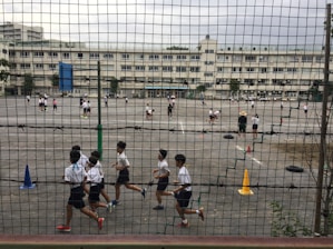 Children are playing and running on a large outdoor playground enclosed by a mesh fence. Various activities are taking place on the asphalt surface, with cones and tires used as part of the setup. Multiple white buildings are visible in the background, indicating a school environment. The area appears spacious and busy, with several groups of students engaging in different games.
