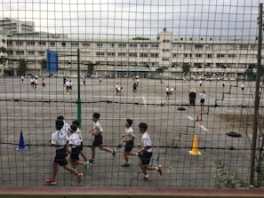 Children are playing and running on a large outdoor playground enclosed by a mesh fence. Various activities are taking place on the asphalt surface, with cones and tires used as part of the setup. Multiple white buildings are visible in the background, indicating a school environment. The area appears spacious and busy, with several groups of students engaging in different games.